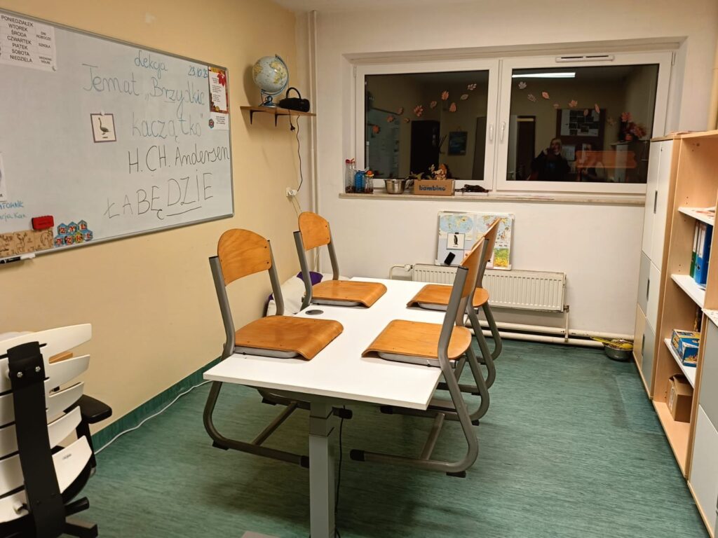 Sala lekcyjna z czteroosobowym stołem, tablicą do nauki i regałami. W pomieszczeniu znajdują się materiały edukacyjne oraz wyposażenie dostosowane do pracy z dziećmi.