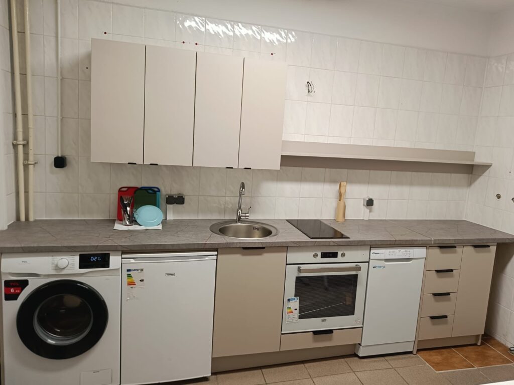 Pomieszczenie kuchenne wyposażone w szafki, zlew, piekarnik, zmywarkę, lodówkę i pralkę. Kuchnia jest jasna i przystosowana do codziennego użytkowania.