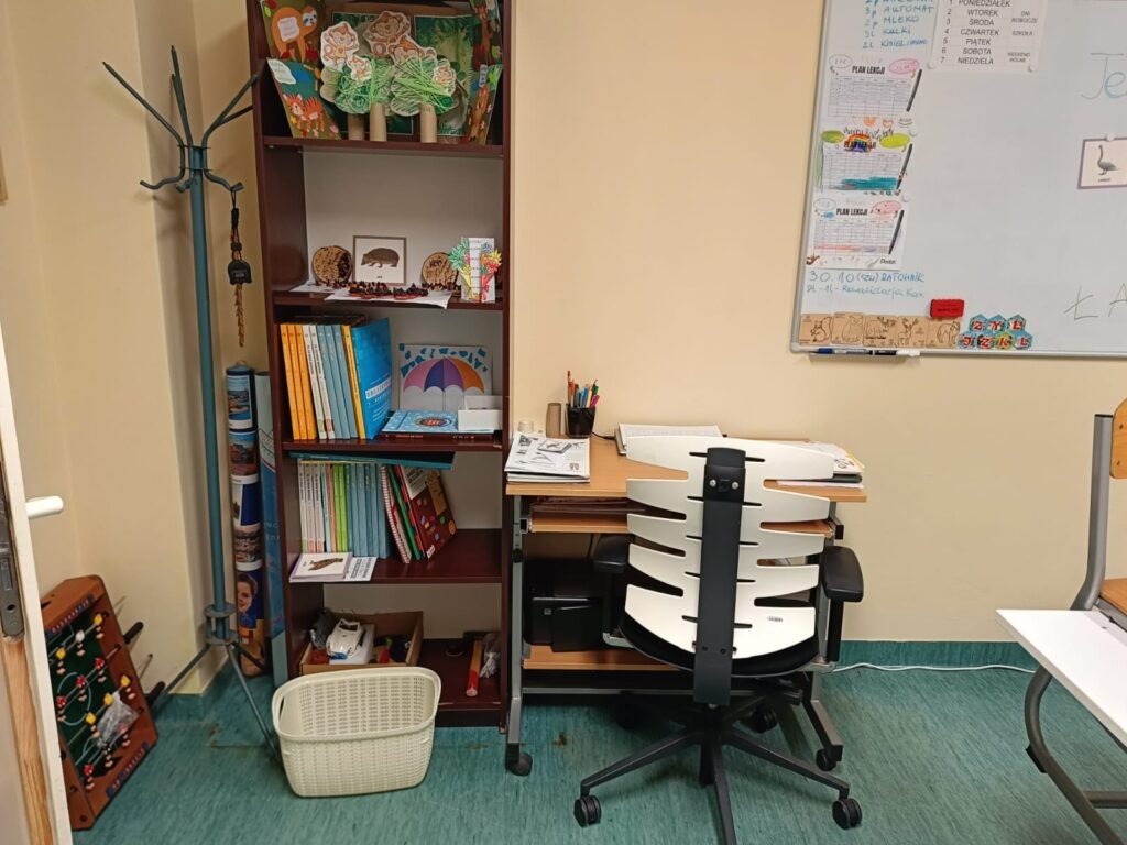 Kącik nauczyciela z biurkiem, regałem z książkami i materiałami edukacyjnymi. Obok znajduje się tablica szkolna oraz wyposażenie sali lekcyjnej.