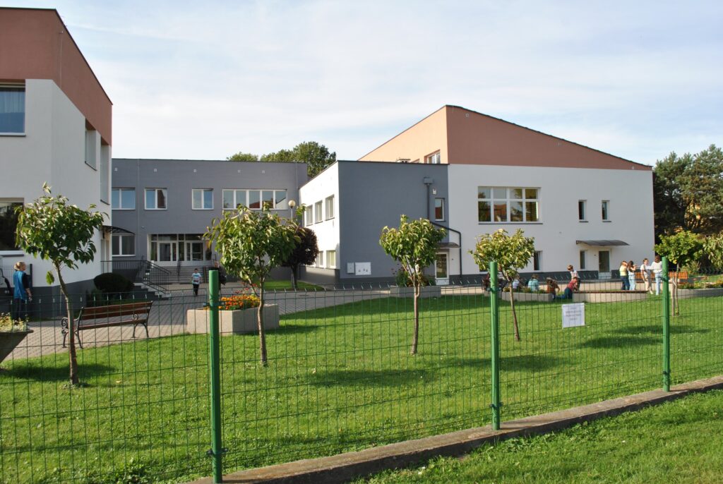 Na obrazku przedstawiony budynek Szkoły Podstawowej im. Jana Pawła II w Smolniku. Jest to budynek szkoły podstawowej z jasną elewacją i dużymi oknami, otoczony zielonym trawnikiem i ogrodzeniem; na terenie widać kilka drzew i dzieci przebywające na zewnątrz.