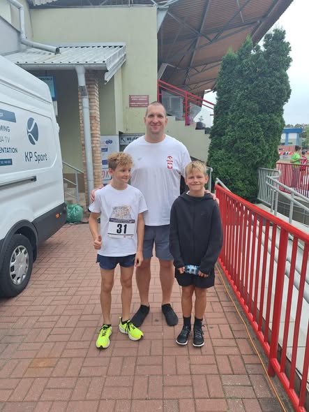 Dwóch chłopców i Wojciech Nowicki pozują przy obiekcie sportowym podczas wydarzenia lekkoatletycznego. Jeden z uczniów ma przypięty numer startowy.