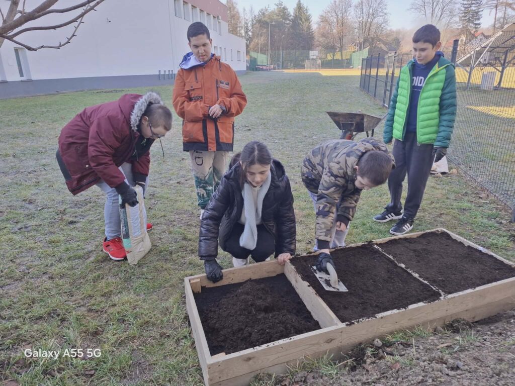 Grupa uczniów przygotowuje ziemię w drewnianych grządkach na terenie szkolnym. Dzieci uczestniczą w zajęciach ogrodniczych na świeżym powietrzu, używając podstawowych narzędzi.