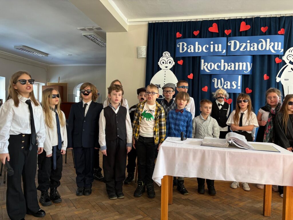 Grupa dzieci występuje podczas uroczystości z okazji Dnia Babci i Dziadka. W tle znajduje się dekoracja z napisem „Babciu, Dziadku, kochamy Was!” oraz czerwone serca. Dzieci stoją przy stole i mają na sobie różnorodne stroje oraz elementy charakteryzacji, między innymi okulary i sztuczne wąsy. Zdjęcie przedstawia szkolny lub przedszkolny występ artystyczny.