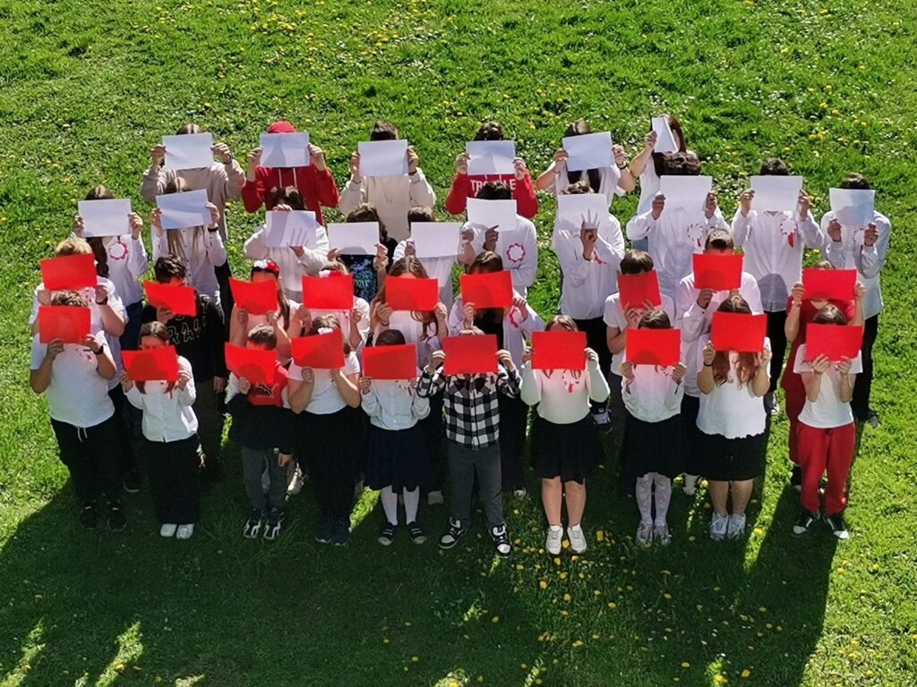 Grupa dzieci stoi na trawie i trzyma przed sobą czerwone oraz białe kartki. Uczestnicy ustawieni są w kilku rzędach na zielonym terenie na świeżym powietrzu. Większość dzieci ma na sobie ubrania w kolorach biało-czerwonych. Zdjęcie zostało wykonane z góry w słoneczny dzień.