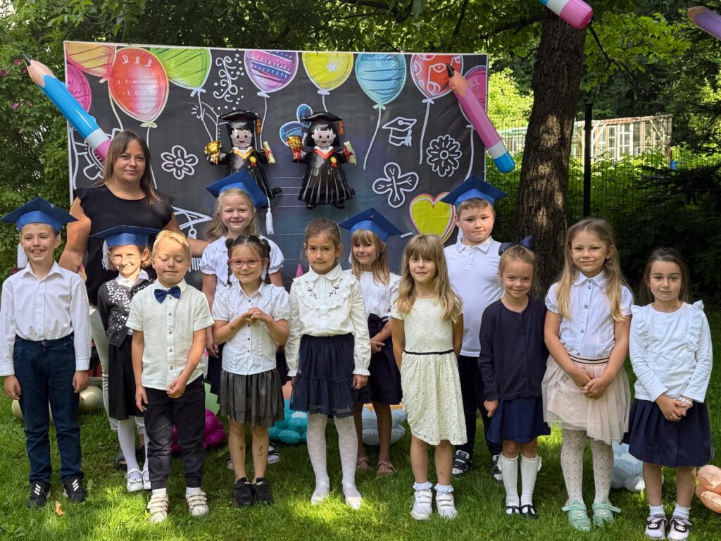 Grupa dzieci wraz z nauczycielką stoi na zewnątrz przed dekoracją związaną z zakończeniem roku lub pasowaniem. Dzieci mają na sobie galowe stroje, a część z nich nosi granatowe birety. W tle widoczna jest kolorowa plansza z balonami i rysunkami. Zdjęcie wykonano w ogrodzie lub na terenie zielonym.