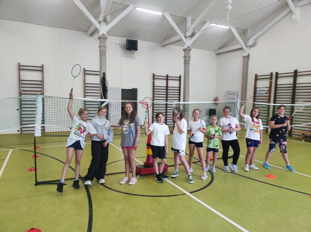 Grupa dzieci stoi w sali gimnastycznej przy siatce do badmintona i trzyma rakietki sportowe. Uczniowie uczestniczą w zajęciach sportowych.