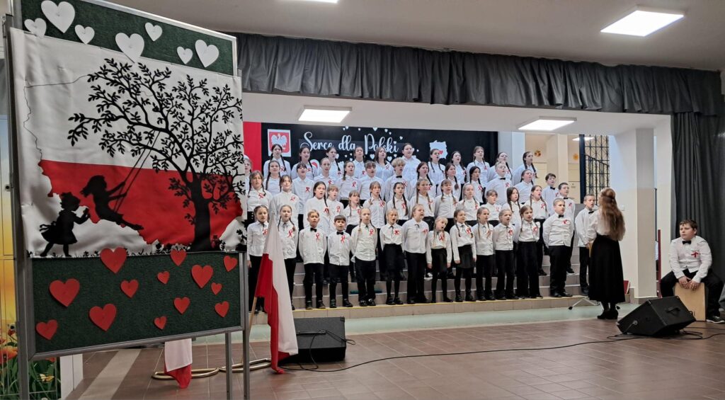 Chór szkolny występuje na scenie podczas uroczystości patriotycznej. Obok widoczna jest dekoracja w barwach biało-czerwonych oraz flaga Polski.