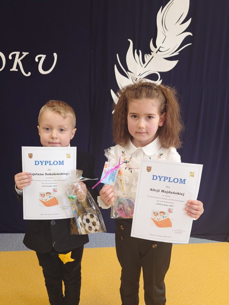 Dwoje małych dzieci stoi przed ciemną dekoracją i trzyma dyplomy oraz upominki. Dzieci ubrane są w eleganckie stroje. Na dyplomach widoczny jest napis „Dyplom”. Zdjęcie przedstawia uczestników konkursu lub szkolnej uroczystości.
