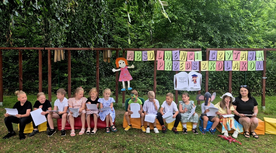 Dzieci siedzą na kolorowych siedziskach na świeżym powietrzu i czytają kartki lub książki. Nad grupą znajduje się dekoracja z napisem „Absolwenci czytają przedszkolakom”. Obok dzieci siedzi dorosła kobieta. W tle widoczne są drzewa i zielona roślinność.