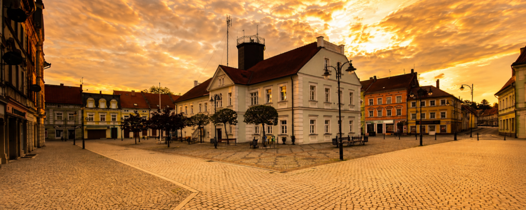 Panoramiczne zdjęcie przedstawia rynek w miejscowości Leśna o zachodzie słońca. W centrum znajduje się jasny, dwukondygnacyjny budynek ratusza z czerwonym dachem oraz niewielką wieżą. Plac wyłożony jest jasną kostką brukową i jest całkowicie pusty – nie ma ludzi ani samochodów.Wokół rynku stoją kolorowe kamienice w odcieniach żółci, pomarańczu i zieleni. Przy budynkach rozmieszczone są stylowe latarnie uliczne oraz niewielkie drzewa o przyciętych koronach.Niebo zajmuje dużą część kadru i ma intensywne barwy złota, pomarańczu i jasnego różu. Chmury są rozświetlone promieniami zachodzącego słońca, co nadaje całej scenie ciepły i spokojny charakter. Zdjęcie sprawia wrażenie bardzo cichego i uporządkowanego miejsca.