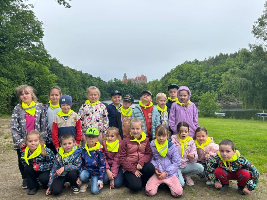 Grupa dzieci stoi na ścieżce w otoczeniu zieleni podczas wycieczki plenerowej. Uczestnicy mają na szyjach jaskrawe chusty odblaskowe i ubrania odpowiednie do chłodniejszej pogody. W tle widoczne są drzewa, jezioro oraz Zamek Czocha położony na wzgórzu. Zdjęcie wykonano w pochmurny dzień.