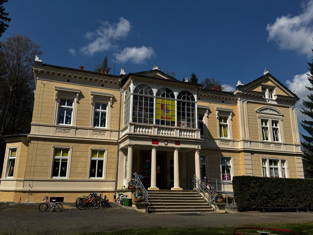 Na zdjęciu widoczny jest zabytkowy budynek placówki edukacyjnej - Przedszkola w Leśnej o jasnej elewacji. Budynek jest dwupiętrowy i ma symetryczną fasadę z dużymi oknami oraz ozdobnymi detalami architektonicznymi. W centralnej części znajduje się przeszklony balkon lub weranda nad głównym wejściem wspartym na kolumnach. Do wejścia prowadzą schody wyposażone w poręcze oraz podjazd dla osób z ograniczoną mobilnością. Przed budynkiem stoją rowery. W tle widoczne są drzewa, a nad budynkiem błękitne niebo z niewielkimi chmurami.