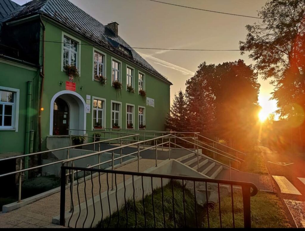Na zdjęciu widoczny jest zielony budynek szkoły sfotografowany o zachodzie słońca. Przy wejściu do budynku znajdują się schody oraz podjazd z metalowymi poręczami dostosowany do potrzeb osób z ograniczoną mobilnością. Na elewacji widoczne są okna z donicami kwiatów oraz tablice informacyjne. Po prawej stronie zdjęcia widać drzewa i nisko położone słońce oświetlające teren wokół budynku. Przed budynkiem znajduje się chodnik oraz ogrodzenie.