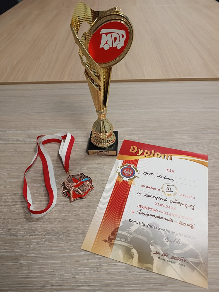 Na pierwszym zdjęciu widoczny jest złoty puchar, medal na biało-czerwonej wstążce oraz dyplom za udział w zawodach młodzieżowych drużyn pożarniczych.
