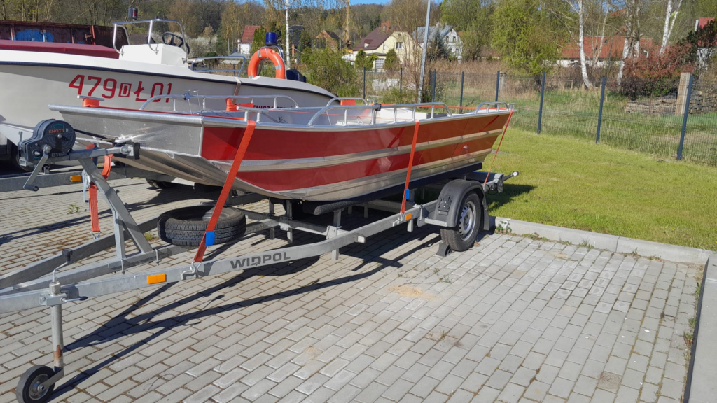 Zdjęcie przedstawia niewielką aluminiową łódź ratowniczą w kolorze czerwono-srebrnym, ustawioną na przyczepie transportowej. Łódź posiada metalowe relingi zabezpieczające oraz płaskie wnętrze umożliwiające przewóz sprzętu lub osób.