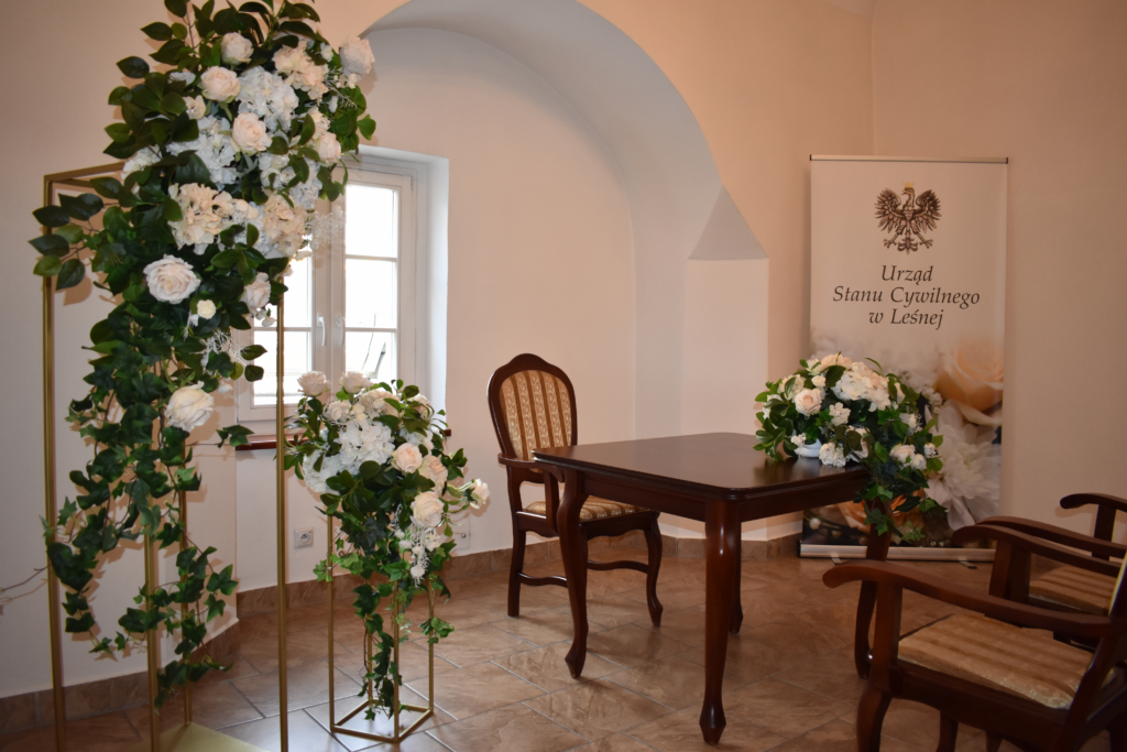 Sala Urzędu Stanu Cywilnego w Leśnej przygotowana do ceremonii ślubnej - drewniany stół i krzesła otoczone dekoracjami z białych, kremowych kwiatów oraz zieleni.